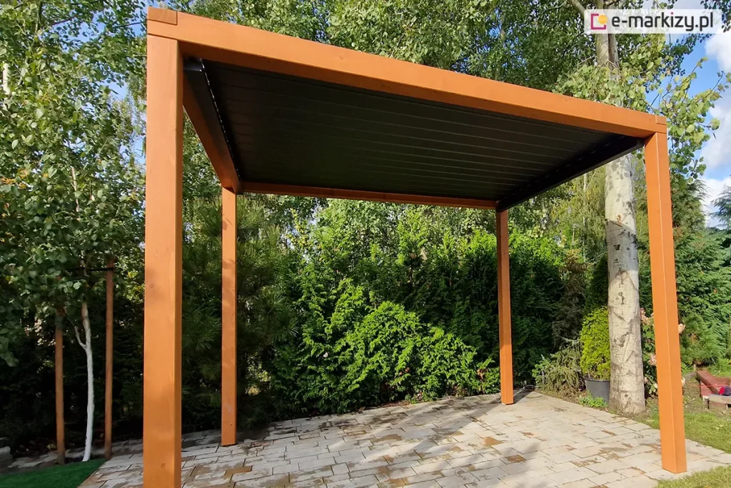 Zdjęcie prezentuje wolnostojącą pergolę ogrodową o solidnej konstrukcji wykonanej z naturalnego drewna w ciepłym odcieniu. Konstrukcja opiera się na czterech kwadratowych słupach nośnych, które podtrzymują prostokątną ramę dachu. Wewnątrz drewnianego szkieletu zamontowano nowoczesny system zadaszenia lamelowego SB400 MD Selt, w kontrastującym, ciemnym kolorze antracytowym. Aluminiowe lamele znajdują się w pozycji całkowicie zamkniętej, tworząc jednolitą i szczelną płaszczyznę chroniącą przestrzeń pod pergolą. Podłoże pod konstrukcją zostało wyłożone jasnymi, prostokątnymi płytami tarasowymi o nieregularnej teksturze. Pergola jest usytuowana w bogato zazielenionej części ogrodu, w bezpośrednim sąsiedztwie gęstych krzewów iglastych oraz wysokich drzew liściastych o jasnej korze, co zapewnia prywatność i naturalne tło dla strefy wypoczynku.