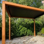 Zdjęcie prezentuje wolnostojącą pergolę ogrodową o solidnej konstrukcji wykonanej z naturalnego drewna w ciepłym odcieniu. Konstrukcja opiera się na czterech kwadratowych słupach nośnych, które podtrzymują prostokątną ramę dachu. Wewnątrz drewnianego szkieletu zamontowano nowoczesny system zadaszenia lamelowego SB400 MD Selt, w kontrastującym, ciemnym kolorze antracytowym. Aluminiowe lamele znajdują się w pozycji całkowicie zamkniętej, tworząc jednolitą i szczelną płaszczyznę chroniącą przestrzeń pod pergolą. Podłoże pod konstrukcją zostało wyłożone jasnymi, prostokątnymi płytami tarasowymi o nieregularnej teksturze. Pergola jest usytuowana w bogato zazielenionej części ogrodu, w bezpośrednim sąsiedztwie gęstych krzewów iglastych oraz wysokich drzew liściastych o jasnej korze, co zapewnia prywatność i naturalne tło dla strefy wypoczynku.