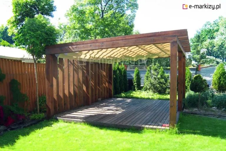 Zdjęcie przedstawia wolnostojącą konstrukcję ogrodową wykonaną z masywnych belek drewnianych o ciemnobrązowym wybarwieniu, pełniącą funkcję zadaszenia tarasu. Wewnątrz konstrukcji, pod górnymi belkami, zamontowano poziomy dach rzymski Supro w kolorze jasnokremowym. Tkanina zadaszenia jest w pełni rozłożona i ułożona w gęste, regularne fale. Konstrukcja posiada jedną ścianę boczną wypełnioną pionowymi, szerokimi deskami, które tworzą ażurową osłonę. Taras jest posadowiony bezpośrednio na trawniku w nasłonecznionej części ogrodu. W bliskim otoczeniu widoczne są gęste nasadzenia z tuj, iglaków oraz drzew liściastych, a po lewej stronie znajduje się wysoki drewniany płot lamelowy. Światło słoneczne przenika przez szczeliny konstrukcji, podkreślając teksturę drewna i tworząc geometryczne wzory cieni na ryflowanych deskach tarasowych.