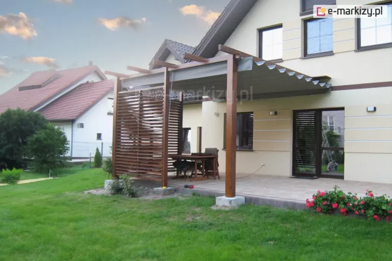 nowoczesna drewniana pergola przyścienna o ciemnobrązowym wybarwieniu, zamontowaną na tarasie domu jednorodzinnego. Konstrukcja wsparta jest na solidnych słupach osadzonych w betonowych stopach fundamentowych i posiada zintegrowaną ściankę boczną z poziomych, ażurowych lameli drewnianych, zapewniającą prywatność. Pod górnym belkowaniem zainstalowano system dachu rzymskiego Supro w kolorze ciemnoszarym, który jest częściowo rozsunięty, tworząc falistą osłonę nad strefą wypoczynkową. Na jasnej posadzce tarasu pod zadaszeniem znajduje się drewniany stół z krzesłami. Elewacja budynku jest wykończona jasnym tynkiem z boniowaniem, a duże przeszklenia tarasowe posiadają ciemne ramy pasujące do koloru dachu rzymskiego. Przed tarasem rozciąga się zadbany trawnik, a w dolnym prawym rogu widoczny jest fragment rabaty z czerwonymi kwiatami. W tle widoczne są sąsiednie budynki mieszkalne z dachami krytymi czerwoną dachówką pod niebem w czasie zmierzchu.