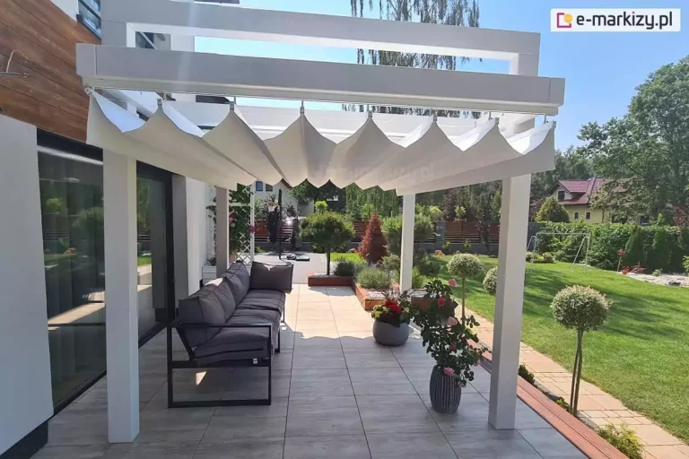 Biała pergola drewniana kolumnowa Moza z opcjonalnym dachem rzymskim Supro w kolorze białym ustawiona na tarasie z meblami oraz roślinami w donicach, w tle widoczny ogród pełen zieleni.