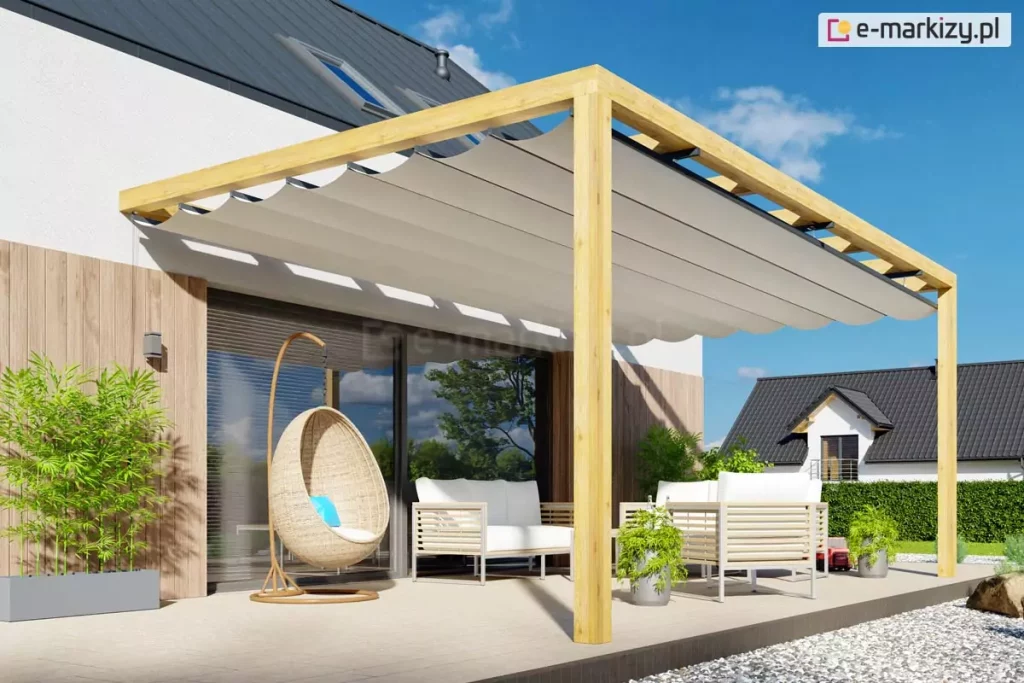 Pergola z tkaniną falowaną ustawiona na prostym, jasnym tarasie przy podwórku wyłożonym kamykami. Na tarasie wyłożonym płytkami ustawiono komplet wypoczynkowy w odcieniach jasnego beżu i bieli oraz fotel typu kokon wykonany z wikliny. Po lewej stronie ustawiono wąską jasnoszarą donicę z zielonymi roślinami. Przy meblach dwie mniejsze doniczki z roślinkami. W tle widoczny żywopłot oraz sąsiedni budynek, a nad wszystkim jasne, niebieskie niebo.