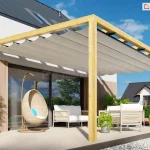 Pergola z tkaniną falowaną ustawiona na prostym, jasnym tarasie przy podwórku wyłożonym kamykami. Na tarasie wyłożonym płytkami ustawiono komplet wypoczynkowy w odcieniach jasnego beżu i bieli oraz fotel typu kokon wykonany z wikliny. Po lewej stronie ustawiono wąską jasnoszarą donicę z zielonymi roślinami. Przy meblach dwie mniejsze doniczki z roślinkami. W tle widoczny żywopłot oraz sąsiedni budynek, a nad wszystkim jasne, niebieskie niebo.