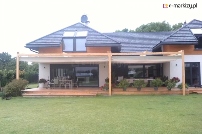 dom jednorodzinny o nowoczesnej bryle z rozłożystym, drewnianym tarasem, nad którym zainstalowano system zadaszenia rzymskiego Silva. Drewniana konstrukcja pergoli, wykonana z jasnych belek, wspiera białe, tekstylne poszycie dachowe, które jest w pełni rozciągnięte, tworząc szeroką strefę cienia nad miejscem wypoczynku. Taras wykonany z desek o ciepłym odcieniu drewna jest wyposażony w duży stół jadalniany z krzesłami oraz liczne rośliny w donicach ustawione wzdłuż krawędzi stopni. Budynek posiada elewację łączącą biały tynk z drewnianą oblicówką oraz ciemnoszarą dachówkę z wbudowanymi oknami połaciowymi. Przed tarasem rozciąga się zadbany, zielony trawnik, a w tle widoczne są korony drzew. Całość kompozycji ukazuje funkcjonalną aranżację przestrzeni ogrodowej, chronioną przed nadmiernym nasłonecznieniem dzięki segmentowemu zadaszeniu.