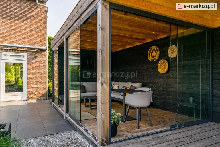 Stylowy ogród letni o konstrukcji hybrydowej, łączącej naturalne drewniane belki konstrukcyjne z nowoczesnym systemem bezramowych szklanych ścian przesuwnych Auris. Zdjęcie przedstawia narożną zabudowę tarasu z widocznym drewnianym stropem i belkami nośnymi. Ściany boczne wykonane są z tafli szkła hartowanego osadzonych w dyskretnych, antracytowych profilach dolnych. Wnętrze urządzone w stylu nowoczesnym posiada czarne ściany z desek, szarą sofę narożną, stół z nowoczesnym krzesłem oraz dekoracyjne plecione talerze na ścianie. Podłoga tarasu wykonana jest z naturalnych desek drewnianych. Zabudowa sąsiaduje z domem z czerwonej cegły i patio wyłożonym dużymi szarymi płytami.