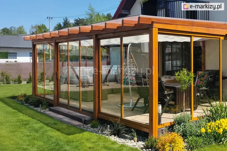 Drewniana pergola o intensywnym, pomarańczowo-brązowym odcieniu, w której zamontowano system przeszkleń Sotro. Przesuwne panele szklane w ramach dopasowanych kolorystycznie do drewna wypełniają całą wysokość konstrukcji. Pod dachem pergoli rozpięta jest biała tkanina osłonowa w formie rzymskiej. Wewnątrz widać strefę wypoczynkową z wiszącym fotelem typu kokon oraz stolikiem kawowym. Całość posadowiona jest na drewnianym podeście otoczonym pasem białego żwiru i kwitnącą roślinnością ogrodową.