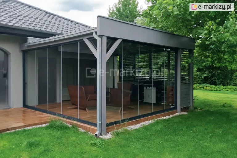 Nowoczesna zabudowa tarasu wykonana w systemie przesuwnych szyb bezramowych Miros, przylegająca do bocznej ściany domu jednorodzinnego z dachem pokrytym dachówką w kolorze grafitowym. Konstrukcja zabudowy oparta na szarej, aluminiowej ramie z pojedynczym słupem narożnym i zintegrowanym systemem rynnowym. Dach zabudowy płaski, jednospadowy, pokryty panelem aluminiowym. Przestrzeń osłonięta szklanymi panelami z dwóch stron, zamontowanymi w dolnych i górnych prowadnicach. Wewnątrz tarasu znajduje się komplet wypoczynkowy z dwoma fotelami i stolikiem kawowym z drewna oraz grill gazowy w czarno-srebrnej obudowie, ustawiony w narożniku przy ścianie z poziomymi aluminiowymi żaluzjami zewnętrznymi. Podłoga zabudowy wyłożona jest deską tarasową o ciepłym, naturalnym odcieniu, z lekkim uniesieniem nad poziomem trawnika. Całość otoczona jest zadbanym, zielonym ogrodem z gęstymi drzewami liściastymi w tle.