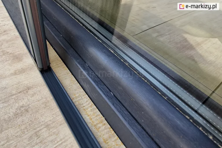 Zbliżenie na ramę okna wykonaną z drewna w kolorze antracytu i ustawioną w jej pobliżu moskitierę plisowaną Top Zag z charakterystycznym niskim progiem, w którego profilu umieszczone są sznurki. Próg moskitiery naklejony na płytkach z drewnopodobnym dekorem.