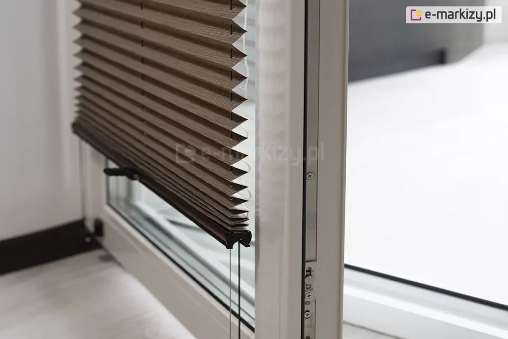 Zdjęcie prezentuje otwarte skrzydło białego okna balkonowego z PCV, na którym zamontowana jest nowoczesna żaluzja plisowana w kolorze ciemnego brązu lub głębokiego taupe. Plisa na okno jest częściowo podciągnięta, a jej materiał układa się w gęste, regularne i ostre załamania tworzące strukturę harmonijki. Dolna krawędź tkaniny wykończona jest smukłym, czarnym profilem sterującym, z którego wystają dwa czarne, dyskretne uchwyty ułatwiające regulację pozycji osłony. Przez środek zagięć materiału oraz przez otwory w dolnej listwie przechodzą pionowe, ciemne sznurki prowadzące, które są napięte i utrzymują plisę równolegle do szyby nawet przy uchylonym oknie. Kadr ukazuje produkt z boku, co pozwala dostrzec techniczną stronę montażu oraz metalowe okucia i uszczelki widoczne na krawędzi otwartego skrzydła okiennego. W tle widać jasne, rozmyte wnętrze oraz fragment podłogi, co podkreśla kontrast między ciemną tkaniną żaluzji a jasną stolarką okienną.
