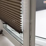 Zdjęcie prezentuje otwarte skrzydło białego okna balkonowego z PCV, na którym zamontowana jest nowoczesna żaluzja plisowana w kolorze ciemnego brązu lub głębokiego taupe. Plisa na okno jest częściowo podciągnięta, a jej materiał układa się w gęste, regularne i ostre załamania tworzące strukturę harmonijki. Dolna krawędź tkaniny wykończona jest smukłym, czarnym profilem sterującym, z którego wystają dwa czarne, dyskretne uchwyty ułatwiające regulację pozycji osłony. Przez środek zagięć materiału oraz przez otwory w dolnej listwie przechodzą pionowe, ciemne sznurki prowadzące, które są napięte i utrzymują plisę równolegle do szyby nawet przy uchylonym oknie. Kadr ukazuje produkt z boku, co pozwala dostrzec techniczną stronę montażu oraz metalowe okucia i uszczelki widoczne na krawędzi otwartego skrzydła okiennego. W tle widać jasne, rozmyte wnętrze oraz fragment podłogi, co podkreśla kontrast między ciemną tkaniną żaluzji a jasną stolarką okienną.