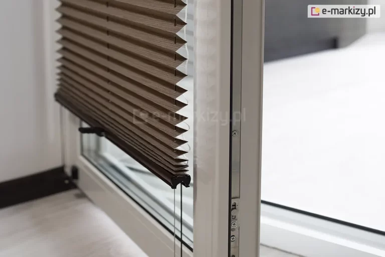 Zdjęcie prezentuje otwarte skrzydło białego okna balkonowego z PCV, na którym zamontowana jest nowoczesna żaluzja plisowana w kolorze ciemnego brązu lub głębokiego taupe. Plisa na okno jest częściowo podciągnięta, a jej materiał układa się w gęste, regularne i ostre załamania tworzące strukturę harmonijki. Dolna krawędź tkaniny wykończona jest smukłym, czarnym profilem sterującym, z którego wystają dwa czarne, dyskretne uchwyty ułatwiające regulację pozycji osłony. Przez środek zagięć materiału oraz przez otwory w dolnej listwie przechodzą pionowe, ciemne sznurki prowadzące, które są napięte i utrzymują plisę równolegle do szyby nawet przy uchylonym oknie. Kadr ukazuje produkt z boku, co pozwala dostrzec techniczną stronę montażu oraz metalowe okucia i uszczelki widoczne na krawędzi otwartego skrzydła okiennego. W tle widać jasne, rozmyte wnętrze oraz fragment podłogi, co podkreśla kontrast między ciemną tkaniną żaluzji a jasną stolarką okienną.