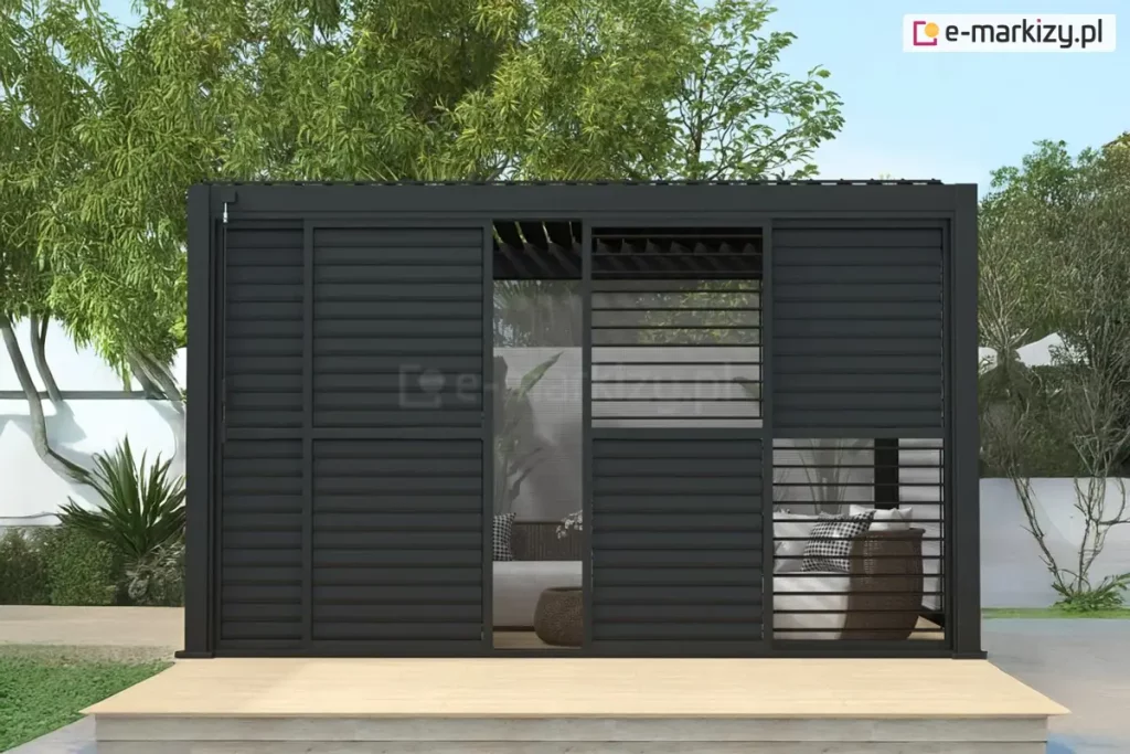 Pergola Gloria ustawiona na drewnianym podeście w ogrodzie z widocznym w tle białym murkiem i drzewami posiada przesłonięte czarnymi panelami lamelowymi przesuwnymi ściany frontowe. Przez uchylone częściowo lamele widoczne ukryte pod pergolą meble wypoczynkowe z technorattanu z białymi siedziskami.