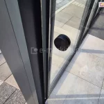 Zbliżenie na detal techniczny zabudowy tarasowej z bezramowym systemem przesuwnym Miros — widoczna czarna, okrągła gałka montowana na tafli szkła hartowanego, służąca jako uchwyt do przesuwania drzwi tarasowych. Szyba umieszczona w dolnej i bocznej prowadnicy wykonanej z antracytowego aluminium, tworzącej stabilną ramę konstrukcyjną. Na podłożu jasne płytki ceramiczne z wyraźnym rysunkiem powierzchni, ułożone równolegle względem ścian. W tle widać zewnętrzne chodniki wyłożone płytkami w różnych odcieniach szarości i beżu, z elementami fakturowanymi i gładkimi, tworzącymi geometryczny wzór. W tafli szkła widoczne odbicia otoczenia oraz gra cieni konstrukcji zadaszenia.