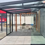 Widok na nowoczesną, całkowicie przeszkloną zabudowę tarasową o czarnej, aluminiowej ramie z prostym dachem z przezroczystych paneli szklanych. Przesuwne, bezramowe szyby Miros przesłaniają przestrzeń z posadzką wyłożoną jasnymi, kwadratowymi płytkami. Panele zsunięte zostały jeden za drugim do prawej strony - do elewacji. Poza zabudową znajdują się dwie prostokątne donice z małą rośliną ozdobną oraz betonowa ławka. W głębi widoczna pergola z drewnianymi słupkami i rozwiniętym, falowanym zadaszeniem materiałowym. Po prawej stronie zdjęcia fragment elewacji z wykończeniem z szarych płyt ceramicznych o strukturze kamienia i pionową, czarną zabudową maskującą. W tle z lewej strony inne elementy ekspozycyjne oraz czerwona tablica reklamowa z białym napisem. Całość znajduje się na terenie wystawowym lub przy punkcie sprzedaży.