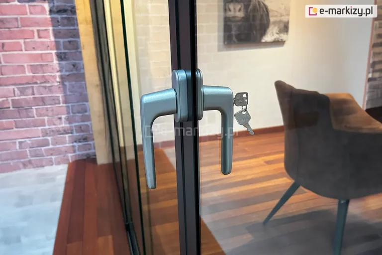 Szczegółowy widok dwustronnej klamki z zamkiem, zamontowanej na pionowym profilu aluminiowym systemu szklanych drzwi przesuwnych Auris. Elementy okuć wykonane są z metalu o satynowym, srebrnym wykończeniu i posiadają nowoczesny, kanciasty kształt z lekko zaokrąglonymi krawędziami. W bębenku zamka po wewnętrznej stronie znajduje się włożony pęk dwóch kluczy na metalowym kółku. Ciemny profil konstrukcyjny oddziela taflę szkła, przez którą widać wnętrze pomieszczenia z drewnianą podłogą ułożoną w jodełkę, brązowym fotelem na cienkich nogach oraz jasną ścianą z ceglanym akcentem po lewej stronie. Na ścianie w tle zawieszony jest obraz przedstawiający zwierzę, a cała scena oświetlona jest miękkim światłem, co podkreśla teksturę materiałów i detale techniczne mechanizmu zamykającego.