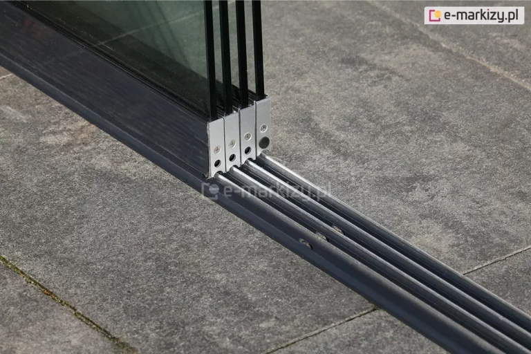 Cztery panele szklanych ścian przesuwnych Auris zsunięte do lewego boku. Pod nimi widoczna dolna prowadnica z czterema torami zamocowana do betonowych płytek podłoża.