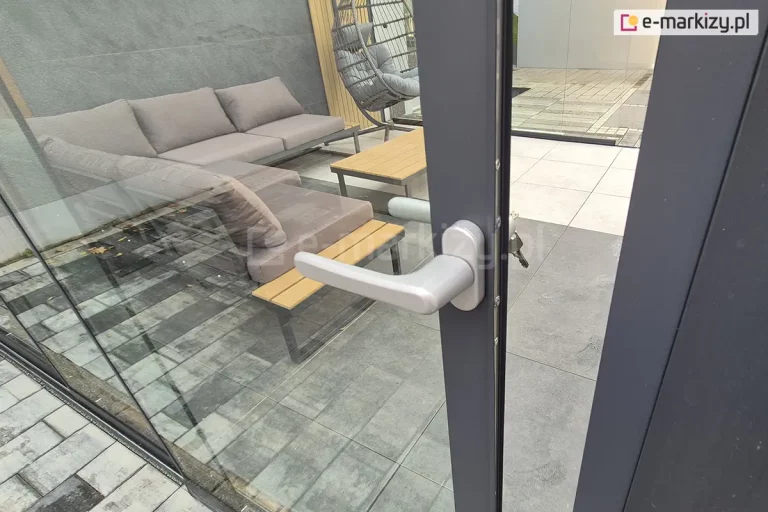 Drzwi przesuwne systemu Auris wyposażone w zamek z kluczem. Na panelu szklanym widoczna jest aluminiowa listwa zamka zamocowana na krawędzi drzwi, wraz z cylindrycznym wkładem i włożonym kluczem. Zamek blokuje drzwi w pozycji zamkniętej, zapewniając zabezpieczenie przestrzeni tarasowej. Tafla szkła jest przezroczysta, zamontowana w aluminiowej prowadnicy u dołu, a całość utrzymana jest w minimalistycznej stylistyce charakterystycznej dla systemu Auris. Ujęcie podkreśla funkcję bezpieczeństwa i wygodę użytkowania szklanych drzwi przesuwnych. W tle pod dachem ze szkła widoczne meble wypoczynkowe, niska sofa oraz stolik z drewnianym blatem i ustawiony w narożniku fotel typu kokon.