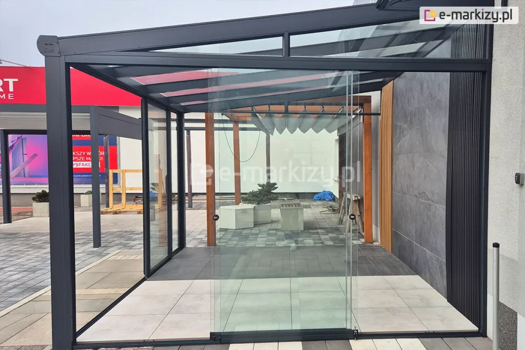 Przeszklona zabudowa tarasu w systemie bezramowym, zbudowana na prostokątnej, czarnej, aluminiowej konstrukcji z dachem z przezroczystego szkła hartowanego. Przesuwne panele szklane zamontowane są w dolnych i górnych prowadnicach, tworząc nowoczesną osłonę tarasu. Panele szklanych ścian przesuwnych Miros zostały zsunięte na środku, jeden za drugim. Na posadzce wewnątrz widoczne są płytki ceramiczne w jasnym odcieniu, a poza konstrukcją zastosowano ciemniejsze płytki ułożone w geometryczny wzór. W centralnej części kadru umieszczone są dwie donice z roślinnością oraz betonowa ławka. Dalej, w tle, zamontowana pergola z materiałowym zadaszeniem w formie zwiniętych fal oraz drewniane elementy konstrukcyjne. Po prawej stronie zdjęcia widoczna elewacja budynku pokryta wielkoformatowymi płytami w odcieniu grafitowym, z pionową czarną listwą dekoracyjną. W lewym górnym rogu fragment czerwonego baneru reklamowego z białym tekstem.