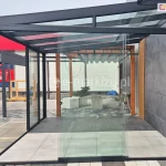 Przeszklona zabudowa tarasu w systemie bezramowym, zbudowana na prostokątnej, czarnej, aluminiowej konstrukcji z dachem z przezroczystego szkła hartowanego. Przesuwne panele szklane zamontowane są w dolnych i górnych prowadnicach, tworząc nowoczesną osłonę tarasu. Panele szklanych ścian przesuwnych Miros zostały zsunięte na środku, jeden za drugim. Na posadzce wewnątrz widoczne są płytki ceramiczne w jasnym odcieniu, a poza konstrukcją zastosowano ciemniejsze płytki ułożone w geometryczny wzór. W centralnej części kadru umieszczone są dwie donice z roślinnością oraz betonowa ławka. Dalej, w tle, zamontowana pergola z materiałowym zadaszeniem w formie zwiniętych fal oraz drewniane elementy konstrukcyjne. Po prawej stronie zdjęcia widoczna elewacja budynku pokryta wielkoformatowymi płytami w odcieniu grafitowym, z pionową czarną listwą dekoracyjną. W lewym górnym rogu fragment czerwonego baneru reklamowego z białym tekstem.