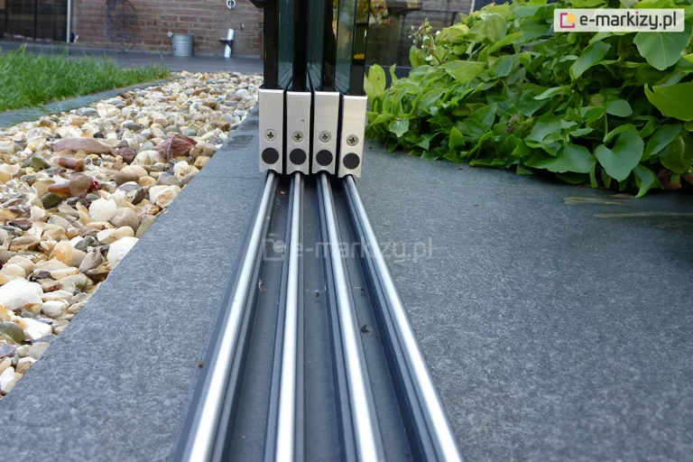 Umieszczona na grafitowej płytce gresowej w pobliżu podłoża wyłożonego kamykami, czterotorowa prowadnica dolna systemu szyb przesuwnych Auris. Na prowadnicy osadzone cztery panele z szybami widoczne od boku. Po prawej stronie paneli duża niska roślinka o zielonych listkach.