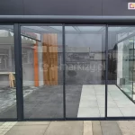 Konstrukcja zadaszenia tarasowego z kompletną zabudową ściany frontowej systemem Sotro. Grafitowa rama konstrukcyjna posiada zaokrąglony profil czołowy, a ściana frontowa składa się z trzech przesuwnych paneli szklanych w cienkich ramkach. Wewnątrz widoczna jest podłoga z jasnych płyt tarasowych oraz dekoracyjna ściana z drewnianych lameli. Zdjęcie wykonane na placu ekspozycyjnym ukazuje transparentność systemu oraz sposób łączenia profili aluminiowych ze szklanym wypełnieniem.