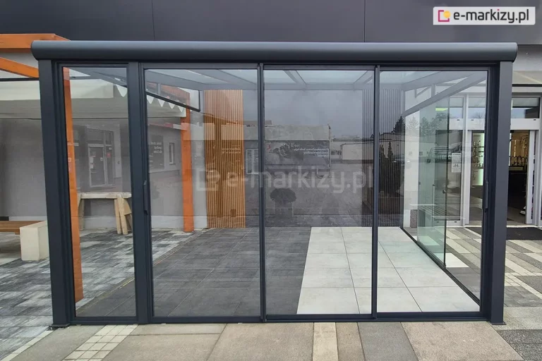 Konstrukcja zadaszenia tarasowego z kompletną zabudową ściany frontowej systemem Sotro. Grafitowa rama konstrukcyjna posiada zaokrąglony profil czołowy, a ściana frontowa składa się z trzech przesuwnych paneli szklanych w cienkich ramkach. Wewnątrz widoczna jest podłoga z jasnych płyt tarasowych oraz dekoracyjna ściana z drewnianych lameli. Zdjęcie wykonane na placu ekspozycyjnym ukazuje transparentność systemu oraz sposób łączenia profili aluminiowych ze szklanym wypełnieniem.