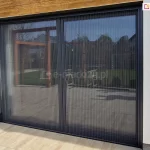 Duże drzwi tarasowe w całości przesłonięte moskitierą plisowaną Top Zag 31 z szarą siatką oraz konstrukcją dopasowaną do barwy ramy okna, zamocowana we wnęce okiennej o białej elewacji oraz na tarasie o jasnych płytkach.