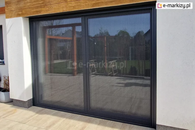 Duże drzwi tarasowe w całości przesłonięte moskitierą plisowaną Top Zag 31 z szarą siatką oraz konstrukcją dopasowaną do barwy ramy okna, zamocowana we wnęce okiennej o białej elewacji oraz na tarasie o jasnych płytkach.