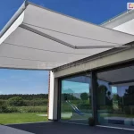 Szeroka, rozłożona osłona tarasowa - markiza Palladio Aluprof w kolorze jasnoszarym, zamontowana nad dużymi przeszkleniami nowoczesnego domu jednorodzinnego. Poszycie wykonane z wysokiej jakości tkaniny jest napięte na dwóch solidnych ramionach przegubowych, które zginają się w połowie swojej długości, stabilizując konstrukcję. Cały system nośny oraz kaseta markizy są polakierowane na kolor jasnoszary, idealnie komponujący się z białą elewacją budynku i antracytowymi ramami okiennymi. Pod osłoną znajduje się ciemny taras, a w dużych szybach przesuwnych drzwi odbija się ogród z zieloną trawą oraz błękitne niebo. W oddali, za niskim ogrodzeniem z siatki, rozciąga się pas gęstego lasu pod czystym niebem. Konstrukcja posiada subtelnie wykończony profil przedni z bocznymi zaślepkami, a całość tworzy geometryczną, minimalistyczną bryłę zintegrowaną z architekturą obiektu.