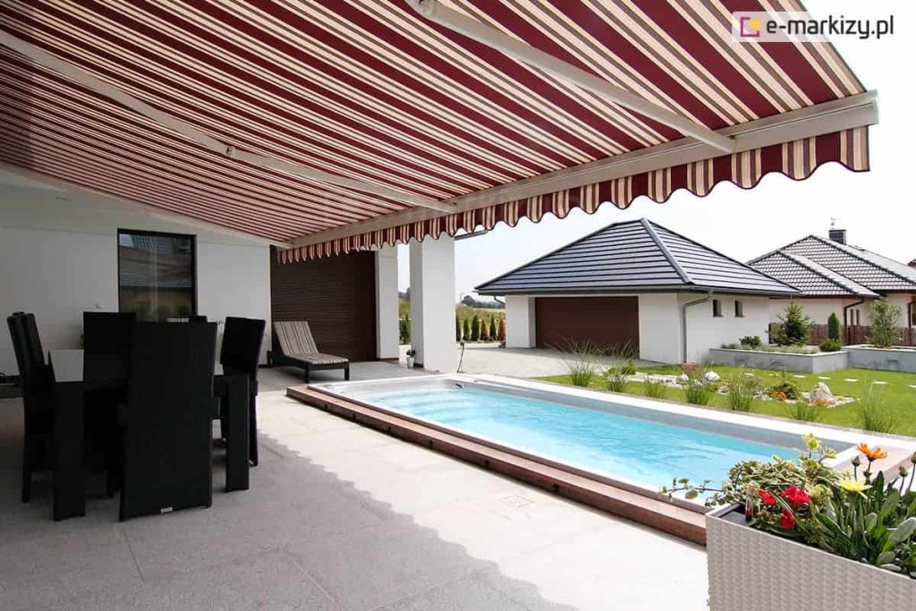 Markiza 7 metrowa Palladio z białą konstrukcją i tkaniną w kasy czerowono-kremowe oraz falbaną o falowanym kształcie osłania taras przy domku, na którym ustawiono czarny zestaw mebli tarasowych oraz białą donicę z kwitnącymi roślinkami. Obok tarasu zainstalowano basen zewnętrzny, za którym widoczny jest zadbany trawnik oraz budynek garażu i domki sąsiadów.