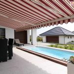 Markiza 7 metrowa Palladio z białą konstrukcją i tkaniną w kasy czerowono-kremowe oraz falbaną o falowanym kształcie osłania taras przy domku, na którym ustawiono czarny zestaw mebli tarasowych oraz białą donicę z kwitnącymi roślinkami. Obok tarasu zainstalowano basen zewnętrzny, za którym widoczny jest zadbany trawnik oraz budynek garażu i domki sąsiadów.