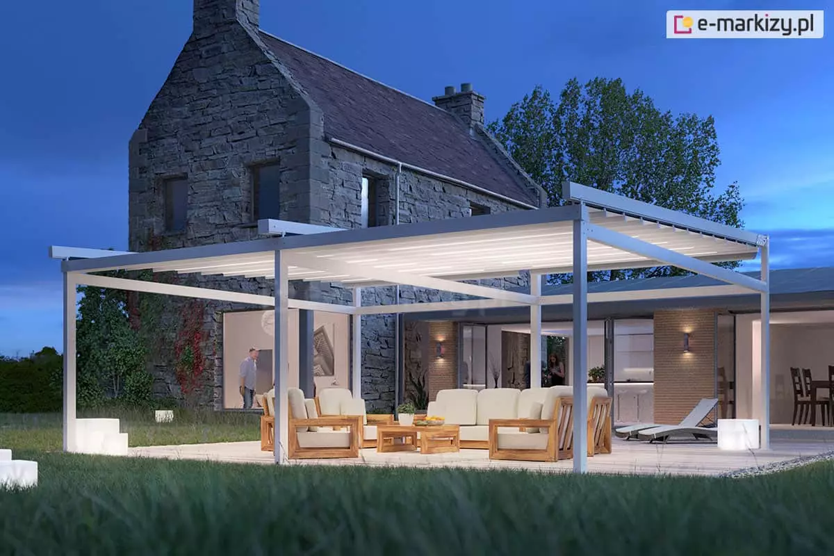 Nowoczesna, wolnostojąca pergola tarasowa Solid w kolorze białym, sfotografowana wieczorem na tle tradycyjnego budynku z szarego kamienia. Konstrukcja posiada zintegrowane oświetlenie LED w formie jasnych pasów na materiale zadaszenia, które emituje intensywne białe światło. Pod pergolą znajduje się luksusowy zestaw mebli ogrodowych z jasnego drewna z grubymi, kremowymi poduszkami oraz niski stolik kawowy. W tle widoczny jest oświetlony dom z dużymi przeszkleniami, przez które widać wnętrze kuchni i jadalni oraz sylwetkę osoby. Na zewnątrz, obok mebli, stoją dwa nowoczesne leżaki oraz świecące białe kostki dekoracyjne rozstawione na trawniku i tarasie. Całość kompozycji dopełnia ciemnoniebieskie, wieczorne niebo i korony drzew w tle.