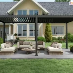 Nowoczesna pergola bioklimatyczna w kolorze antracytowym, zainstalowana jako wolnostojąca konstrukcja na betonowym podłożu przed domem jednorodzinnym. Pergola składa się z dwóch segmentów wspartych na solidnych pionowych słupach, z dachem wykonanym z ruchomych lameli aluminiowych. W lewej części dachu lamele są ustawione pod kątem, ukazując prześwity nieba, natomiast w prawej części są całkowicie zamknięte, tworząc zwartą osłonę. Pod pergolą zaaranżowano strefę wypoczynkową z luksusowymi meblami ogrodowymi na drewnianych podstawach, w tym dwiema sofami i fotelami z jasnymi poduszkami oraz drewnianym stolikiem kawowym w centrum. Po prawej stronie przy słupie znajduje się podłużna, czarna donica z roślinnością. W tle widoczny jest piętrowy dom o żółtej elewacji z licznymi przeszkleniami oraz zadbany trawnik z ozdobnymi krzewami i drzewami iglastymi. Po obu stronach konstrukcji zwisają korby służące do manualnej regulacji kąta nachylenia lameli dachowych.