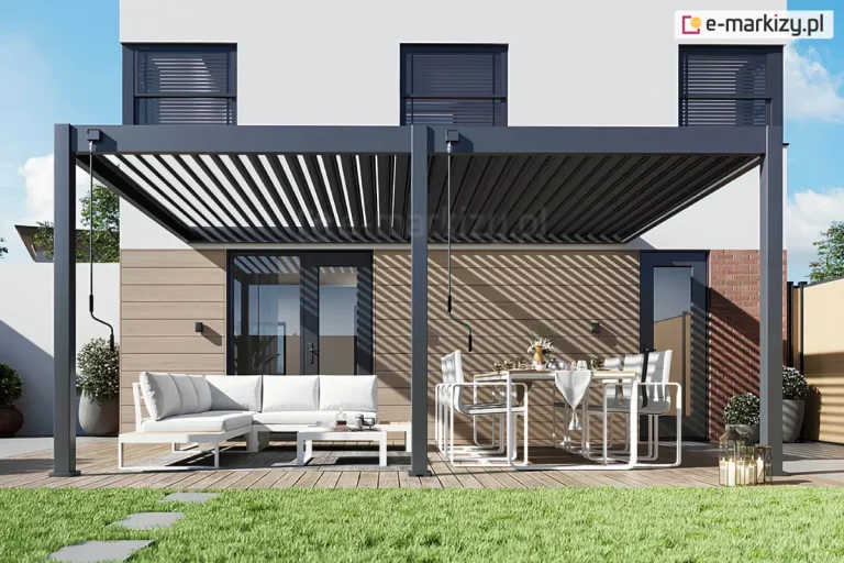 Dwumodułowa pergola tarasowa lamelowa zamontowana przy nowoczesnym budynku mieszkalnym, zaprezentowana w pełnym słońcu. Aluminiowa konstrukcja w kolorze ciemnego antracytu opiera się na trzech pionowych słupach o kwadratowym przekroju, kotwionych do drewnianego tarasu. Zadaszenie składa się z dwóch niezależnych sekcji lameli: lewa sekcja jest otwarta, ukazując błękitne niebo i rzucając wyraźny, pasiasty cień na elewację domu, natomiast prawa sekcja pozostaje przymknięta lub otwarta pod innym kątem. Przy skrajnym lewym słupie oraz przy słupie środkowym zamontowane są manualne korby sterujące z czarnymi uchwytami. Pod pergolą urządzono dwie strefy wypoczynkowe z białymi meblami ogrodowymi: po lewej stronie znajduje się duży narożnik z jasnymi poduszkami i stolik kawowy, a po prawej stronie stół jadalniany z sześcioma krzesłami, na którym ustawiona jest zastawa. Tło stanowi jasna elewacja budynku wykończona częściowo panelami w kolorze drewna oraz cegłą, z widocznymi ciemnymi ramami okiennymi i drzwiami tarasowymi. W dolnej części zdjęcia widoczny jest fragment zielonego trawnika oraz ścieżka z szarych płyt betonowych, a po prawej stronie donice z niską roślinnością i lampiony ustawione na deskach tarasowych.