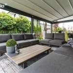 Zdjęcie prezentuje wnętrze nowoczesnej, przestronnej pergoli tarasowej tarasola elegancy, zaaranżowanej jako luksusowa strefa wypoczynkowa. Pod jasnym, segmentowym zadaszeniem tkaninowym znajduje się duży narożny zestaw mebli ogrodowych z grafitowymi poduszkami, ustawiony na szarym tarasie z płyt. Centralnym punktem jest niski stolik kawowy z drewnianym blatem, pod którym rozłożono dekoracyjny dywan zewnętrzny. Przestrzeń pod pergolą tarasola elegancy ozdobiona jest licznymi detalami, takimi jak rzeźby głów Buddy, donice z formowaną zielenią oraz dekoracyjne wazony. Przeszklone ściany boczne pergoli otwierają widok na zadbany ogród z gęstym żywopłotem z tui oraz innymi krzewami ozdobnymi, co zapewnia prywatność przy jednoczesnym zachowaniu kontaktu z naturą. Konstrukcja o ciemnych profilach aluminiowych podkreśla nowoczesny i elegancki charakter całej zabudowy tarasowej.