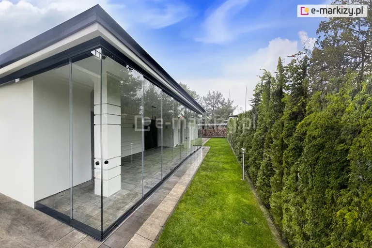 Nowoczesna zabudowa tarasu wykonana z bezramowego systemu przesuwnych szyb ze szkła hartowanego Miros, zamontowana na krawędzi budynku o białej elewacji i ciemnym, płaskim dachu z wyraźnym okapem. Szklane panele osłaniają przestrzeń wykończoną jasnoszarą, prostokątną płytką podłogową ułożoną równolegle do krawędzi domu. Wnętrze zabudowy jest puste, z widocznymi detalami architektonicznymi, takimi jak słup wykończony dekoracyjnym frezowaniem poziomym. Po prawej stronie kadru znajduje się zadbany pas intensywnie zielonego trawnika, wzdłuż którego biegnie wąska ścieżka z kostki brukowej, prowadząca w stronę tylnej części ogrodu. Po prawej stronie ścieżki widoczny jest gęsty żywopłot z wysokich, przystrzyżonych tuj, wzdłuż którego zamontowano pojedyncze niskie lampy ogrodowe. W tle widoczna siatka ogrodzeniowa oraz drzewa liściaste i iglaste, a nad wszystkim niebo z częściowym zachmurzeniem i błękitnymi prześwitami.