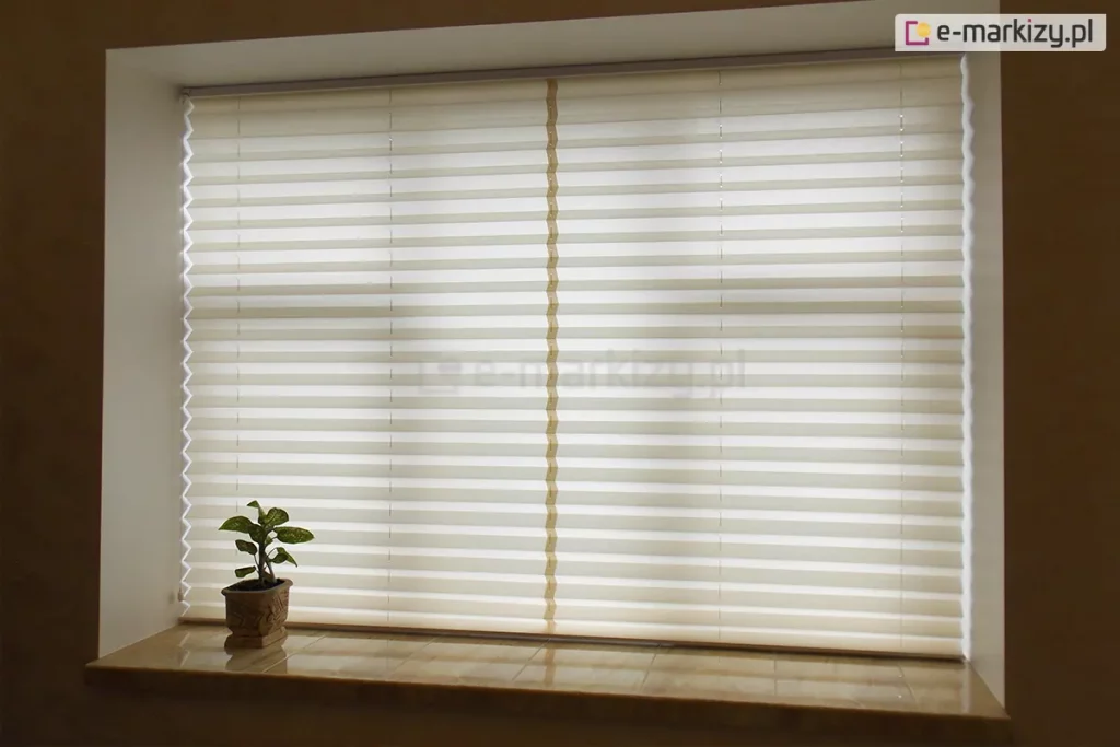 Szerokie ujęcie prezentuje zamontowaną we wnęce okiennej, w pełni rozwiniętą osłonę, którą jest żaluzja plisowana. Plisa na okno w kolorze kremowym lub złamanej bieli szczelnie zasłania całą powierzchnię przeszklenia, wprowadzając do wnętrza miękkie, rozproszone światło dzienne. Materiał żaluzji charakteryzuje się gęstym, poziomym plisowaniem, tworzącym rytmiczny wzór równoległych linii na całej wysokości okna. Konstrukcja jest stabilizowana przez trzy pionowe rzędy sznurków prowadzących – dwa znajdują się blisko bocznych krawędzi, a jeden przebiega dokładnie przez środek tkaniny, co zapewnia idealne napięcie materiału na dużej powierzchni. Osłona jest osadzona w minimalistycznych, jasnych profilach, które niemal zlewają się z ramą okienną. Poniżej okna znajduje się szeroki parapet wykonany z błyszczących kafli lub kamienia w odcieniu beżowym, na którym po lewej stronie ustawiona jest mała roślina doniczkowa w ozdobnej, brązowej doniczce. Całość aranżacji podkreśla funkcjonalność żaluzji w kontrolowaniu prywatności i nasłonecznienia, tworząc przytulną atmosferę w pomieszczeniu.
