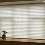 Szerokie ujęcie prezentuje zamontowaną we wnęce okiennej, w pełni rozwiniętą osłonę, którą jest żaluzja plisowana. Plisa na okno w kolorze kremowym lub złamanej bieli szczelnie zasłania całą powierzchnię przeszklenia, wprowadzając do wnętrza miękkie, rozproszone światło dzienne. Materiał żaluzji charakteryzuje się gęstym, poziomym plisowaniem, tworzącym rytmiczny wzór równoległych linii na całej wysokości okna. Konstrukcja jest stabilizowana przez trzy pionowe rzędy sznurków prowadzących – dwa znajdują się blisko bocznych krawędzi, a jeden przebiega dokładnie przez środek tkaniny, co zapewnia idealne napięcie materiału na dużej powierzchni. Osłona jest osadzona w minimalistycznych, jasnych profilach, które niemal zlewają się z ramą okienną. Poniżej okna znajduje się szeroki parapet wykonany z błyszczących kafli lub kamienia w odcieniu beżowym, na którym po lewej stronie ustawiona jest mała roślina doniczkowa w ozdobnej, brązowej doniczce. Całość aranżacji podkreśla funkcjonalność żaluzji w kontrolowaniu prywatności i nasłonecznienia, tworząc przytulną atmosferę w pomieszczeniu.