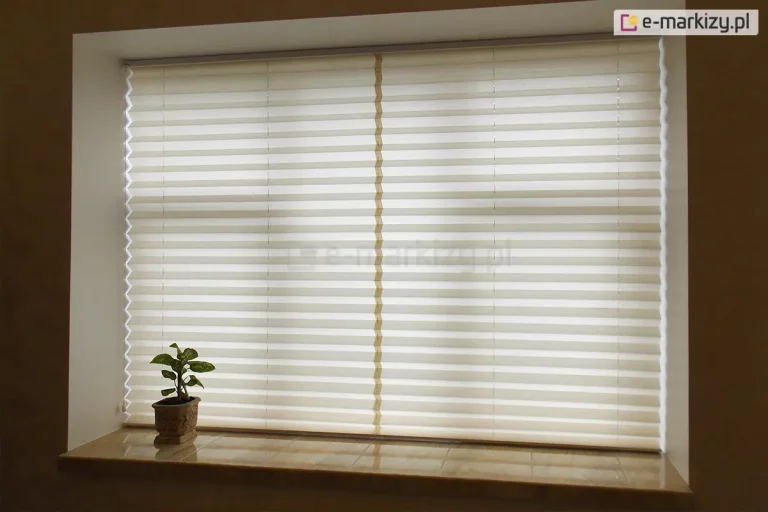 Szerokie ujęcie prezentuje zamontowaną we wnęce okiennej, w pełni rozwiniętą osłonę, którą jest żaluzja plisowana. Plisa na okno w kolorze kremowym lub złamanej bieli szczelnie zasłania całą powierzchnię przeszklenia, wprowadzając do wnętrza miękkie, rozproszone światło dzienne. Materiał żaluzji charakteryzuje się gęstym, poziomym plisowaniem, tworzącym rytmiczny wzór równoległych linii na całej wysokości okna. Konstrukcja jest stabilizowana przez trzy pionowe rzędy sznurków prowadzących – dwa znajdują się blisko bocznych krawędzi, a jeden przebiega dokładnie przez środek tkaniny, co zapewnia idealne napięcie materiału na dużej powierzchni. Osłona jest osadzona w minimalistycznych, jasnych profilach, które niemal zlewają się z ramą okienną. Poniżej okna znajduje się szeroki parapet wykonany z błyszczących kafli lub kamienia w odcieniu beżowym, na którym po lewej stronie ustawiona jest mała roślina doniczkowa w ozdobnej, brązowej doniczce. Całość aranżacji podkreśla funkcjonalność żaluzji w kontrolowaniu prywatności i nasłonecznienia, tworząc przytulną atmosferę w pomieszczeniu.