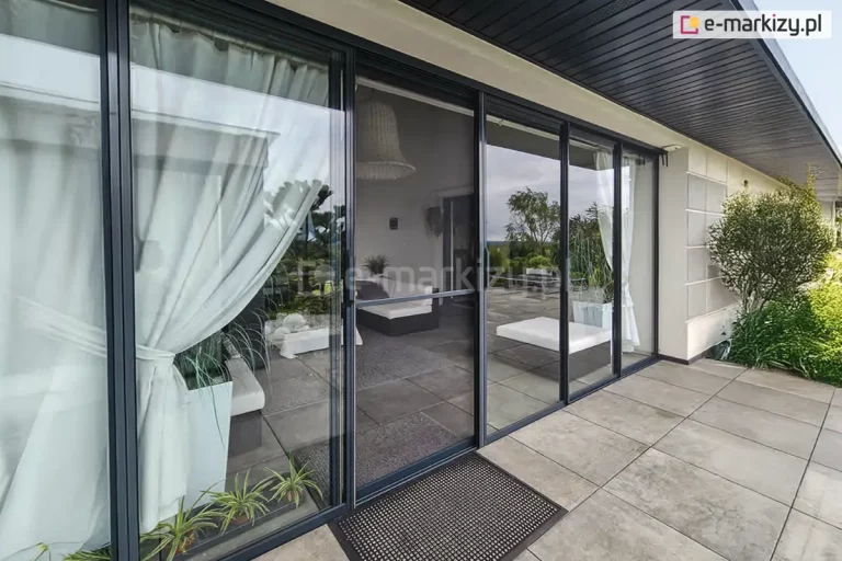 szerokie przeszklenia przesuwne systemu Sotro zamontowane w nowoczesnym budynku z zadaszonym tarasem. Grafitowe ramy okalają duże tafle szkła, z których jedna jest częściowo przysłonięta białą firaną upiętą z boku. Przeszklona ściana prowadzi do wnętrza z szarymi meblami, nad którymi wisi pleciony żyrandol w stylu boho. Taras wyłożony jest dużymi, jasnoszarymi płytami gresowymi, a przed wejściem leży czarna wycieraczka ażurowa. W szybach odbija się zielony ogród oraz błękitne niebo, co podkreśla transparentność i wysoką jakość zastosowanego szklenia.