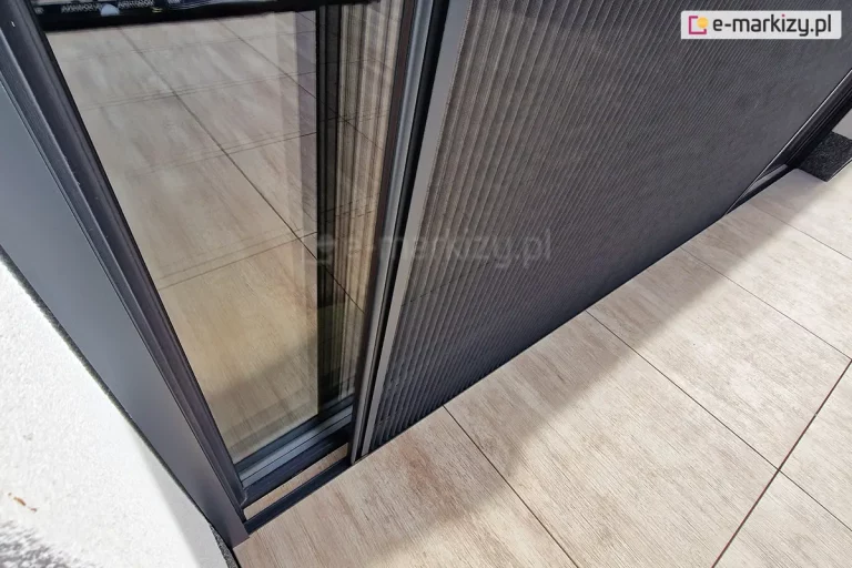 Dwustronnie otwierana moskitiera Top Zag zamontowana na tarasie z dużych płyt o drewnopodobnym wzorze. Ukazany z góry próg dolny, odsunięty od ramy okna, nie utrudnia przechodzenia. Profile moskitiery dopasowano kolorystycznie do barw okna.