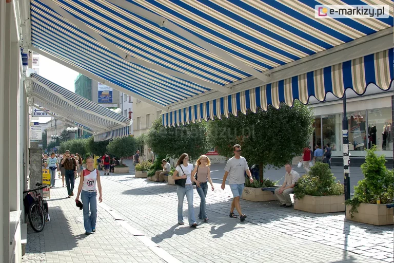 Zdjęcie przedstawia tętniący życiem miejski deptak wyłożony szarą kostką brukową, nad którym zamontowano dwie duże markizy tarasowe. Markizy palladio posiadają charakterystyczne poszycie w szerokie, pionowe pasy w kolorach białym, niebieskim i beżowym, zakończone ozdobną, falistą falbaną o tym samym wzorze. Konstrukcje markiz opierają się na białych ramionach przymocowanych do elewacji budynków, rzucając wyraźny cień na znaczną część chodnika. Wzdłuż traktu pieszego ustawione są jasne, betonowe donice z przyciętymi w kulę krzewami oraz niskimi roślinami ozdobnymi, przy których znajdują się ławki dla przechodniów. Po deptaku spacerują ludzie w letnich ubraniach, a w tle widoczna jest dalsza część ulicy z witrynami sklepowymi, szyldami reklamowymi oraz zaparkowanym przy ścianie budynku rowerem. Światło słoneczne jest intensywne, tworząc wyraźne kontrasty między zacienionymi strefami pod markizami a jasno oświetloną środkową częścią promenady.