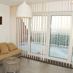 Zdjęcie przedstawia nowoczesny salon z dużym przeszkleniem balkonowym, które osłaniają żaluzje pionowe w jasnym, kremowym kolorze. System składa się z licznych, wąskich lameli zwisających od sufitu aż do samej podłogi, zamontowanych na długiej, białej szynie. Pasy materiału są ustawione prostopadle do okna, co pozwala na pełny wgląd na ośnieżony taras oraz panoramę miasta z widocznymi budynkami mieszkalnymi w oddali. W dolnej części lameli widoczne są obciążniki połączone białym łańcuszkiem kulkowym, który stabilizuje konstrukcję i zapewnia równomierne odstępy między pasami. Wnętrze pokoju jest urządzone w stylu skandynawskim lub boho; po lewej stronie stoi wiklinowa sofa z beżowymi poduszkami oraz pasujący do niej okrągły, pleciony stolik kawowy. Nad meblami wisi duża, biała lampa o kloszu przypominającym plecionkę, a na ścianie umieszczono niewielki, kolorowy obraz w złotej ramie. Podłoga wykonana jest z dużych, jasnoszarych kafli, które odbijają naturalne światło wpadające przez żaluzje pionowe.