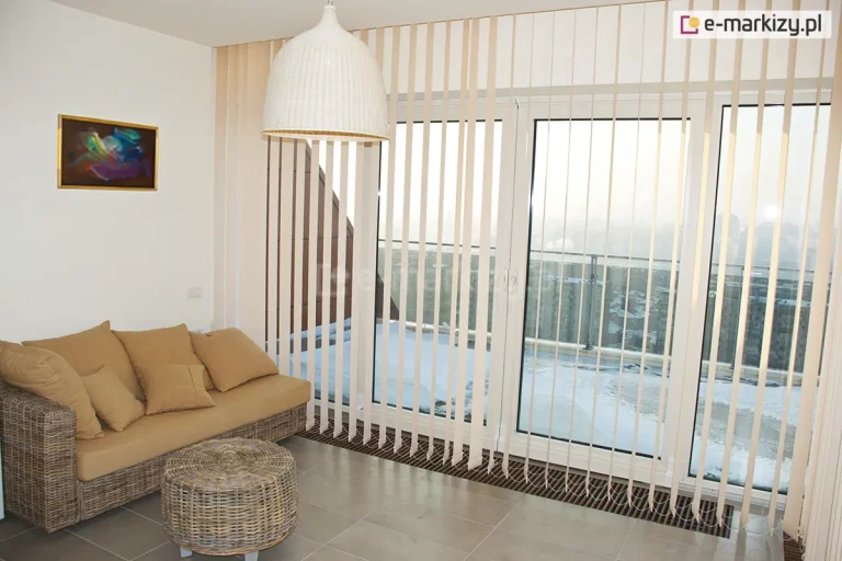 Zdjęcie przedstawia nowoczesny salon z dużym przeszkleniem balkonowym, które osłaniają żaluzje pionowe w jasnym, kremowym kolorze. System składa się z licznych, wąskich lameli zwisających od sufitu aż do samej podłogi, zamontowanych na długiej, białej szynie. Pasy materiału są ustawione prostopadle do okna, co pozwala na pełny wgląd na ośnieżony taras oraz panoramę miasta z widocznymi budynkami mieszkalnymi w oddali. W dolnej części lameli widoczne są obciążniki połączone białym łańcuszkiem kulkowym, który stabilizuje konstrukcję i zapewnia równomierne odstępy między pasami. Wnętrze pokoju jest urządzone w stylu skandynawskim lub boho; po lewej stronie stoi wiklinowa sofa z beżowymi poduszkami oraz pasujący do niej okrągły, pleciony stolik kawowy. Nad meblami wisi duża, biała lampa o kloszu przypominającym plecionkę, a na ścianie umieszczono niewielki, kolorowy obraz w złotej ramie. Podłoga wykonana jest z dużych, jasnoszarych kafli, które odbijają naturalne światło wpadające przez żaluzje pionowe.