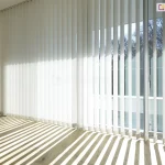 Zdjęcie prezentuje jasne, nowoczesne wnętrze z dużym przeszkleniem, na którym zamontowane zostały żaluzje pionowe w kolorze białym. Osłona składa się z bardzo długich lameli, które sięgają od samego sufitu do jasnej, drewnianej podłogi. Pasy materiału są ustawione w dwóch różnych konfiguracjach: po lewej stronie są całkowicie zamknięte, tworząc jednolitą, białą płaszczyznę, natomiast po prawej stronie zostały ustawione prostopadle do szyb. Takie ustawienie sprawia, że do wnętrza wpada ostre światło słoneczne, które rzuca na podłogę geometryczny wzór szerokich, równoległych cieni. Przez otwarte żaluzje pionowe widać fragment tarasu z białą balustradą oraz zielone korony drzew w tle. W dolnej części przesłony, tuż nad podłogą, widoczny jest system białych łańcuszków kulkowych, które łączą poszczególne lamele, stabilizując ich położenie. Całość kompozycji podkreśla przestronność i minimalistyczny design pomieszczenia, w którym główną rolę gra światło i cień.