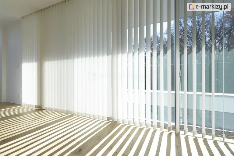 Zdjęcie prezentuje jasne, nowoczesne wnętrze z dużym przeszkleniem, na którym zamontowane zostały żaluzje pionowe w kolorze białym. Osłona składa się z bardzo długich lameli, które sięgają od samego sufitu do jasnej, drewnianej podłogi. Pasy materiału są ustawione w dwóch różnych konfiguracjach: po lewej stronie są całkowicie zamknięte, tworząc jednolitą, białą płaszczyznę, natomiast po prawej stronie zostały ustawione prostopadle do szyb. Takie ustawienie sprawia, że do wnętrza wpada ostre światło słoneczne, które rzuca na podłogę geometryczny wzór szerokich, równoległych cieni. Przez otwarte żaluzje pionowe widać fragment tarasu z białą balustradą oraz zielone korony drzew w tle. W dolnej części przesłony, tuż nad podłogą, widoczny jest system białych łańcuszków kulkowych, które łączą poszczególne lamele, stabilizując ich położenie. Całość kompozycji podkreśla przestronność i minimalistyczny design pomieszczenia, w którym główną rolę gra światło i cień.