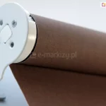 Zbliżenie łączenia mechanizmu rolety wolnowiszącej w kolorze białym umieszczonego w białym uchwycie ściennym rolety Plus. Obok widoczna także tkanina rolety w odcieniu mlecznej czekolady.