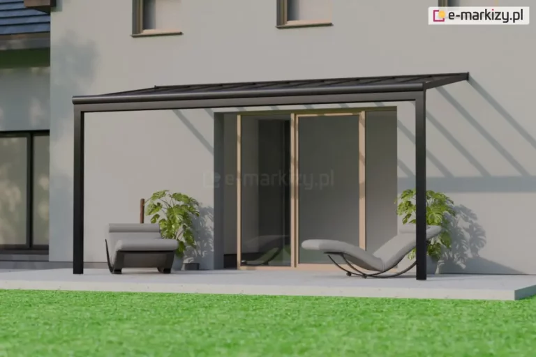 Nowoczesny taras przy jasnoszarej ścianie domu, z zamontowanym czarnym, prostokątnym zadaszeniem z przezroczystych paneli, przepuszczających światło i rzucających wyraźne cienie belek na elewację. Pod zadaszeniem, na jasnym, gładkim podłożu, stoją dwa identyczne leżaki w nowoczesnym stylu — szare z wygiętą konstrukcją i czarną, zaokrągloną podstawą typu kołyskowego. Jeden leżak znajduje się po lewej stronie tarasu i ustawiony jest frontalnie, natomiast drugi, po prawej stronie, ułożony jest bokiem, z widocznym profilem siedziska i podpórkami. Za każdym z leżaków stoi duża donica z zieloną rośliną ozdobną o dużych, gęstych liściach. W tle widoczne są przesuwne drzwi tarasowe w jasnobrązowej, drewnianej ramie, prowadzące do wnętrza budynku. Po lewej stronie kadru częściowo widoczny jest inny fragment domu z dużym oknem i ciemnoniebieskim dachem. Przed tarasem znajduje się intensywnie zielony trawnik.