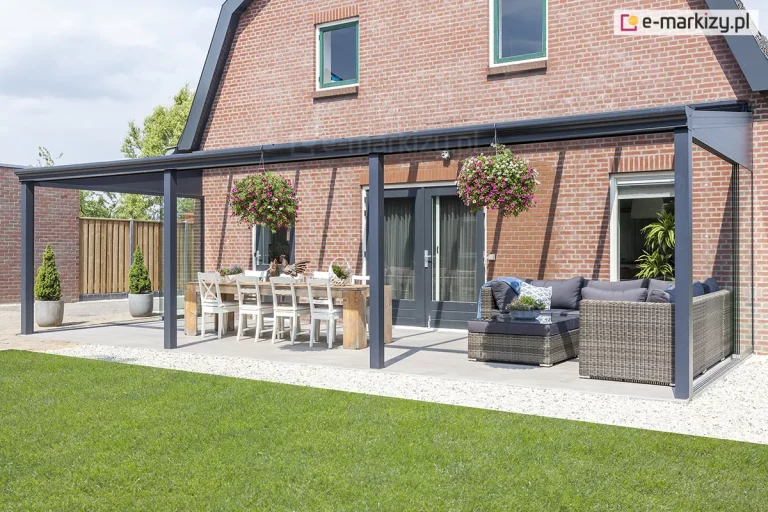 Szerokie zadaszenie tarasowe przy budynku z czerwonej cegły, wykonane z antracytowych profili aluminiowych. Konstrukcja dachu jest przeszklona, a boki tarasu zabezpieczono stałymi ściankami szklanymi w kształcie trapezów oraz przesuwnymi panelami. Pod zadaszeniem znajduje się duży drewniany stół z białymi krzesłami oraz narożny zestaw wypoczynkowy z technorattanu z szarymi poduszkami. Z konstrukcji zwisają donice z kwitnącymi kwiatami (surfinie). Podłoże tarasu jest jasne i gładkie, otoczone opaską z białego kamienia ozdobnego i trawnikiem.