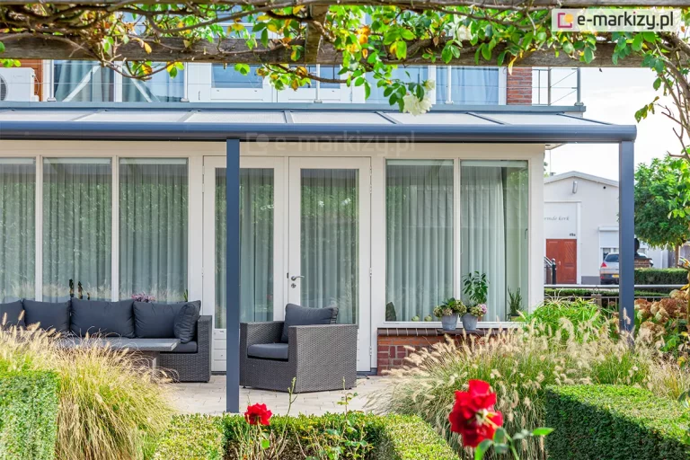 Szerokie, nowoczesne zadaszenie tarasowe Lunaro o konstrukcji w kolorze ciemnego antracytu, zamontowane do białej ściany budynku z dużą ilością przeszkleń. Dach o niskim kącie nachylenia posiada wypełnienie z półprzezroczystych paneli poliwęglanowych, które są podtrzymywane przez smukłe profile i oparte na trzech słupach nośnych usytuowanych w jednej linii. Pod zadaszeniem, na jasnym tarasie, zaaranżowano strefę wypoczynkową z meblami z ciemnego technorattanu, w tym długą sofę i fotel z szarymi poduszkami. Tło stanowią białe drzwi tarasowe oraz wysokie okna z jasnymi zasłonami, a na parapecie ustawiono doniczki z kwiatami. Pierwszy plan oraz otoczenie tarasu wypełnia bujna roślinność, w tym ozdobne trawy, niskie krzewy oraz czerwone róże, a całość ujęcia jest kadrowana przez gałęzie pnączy zwisające z góry.